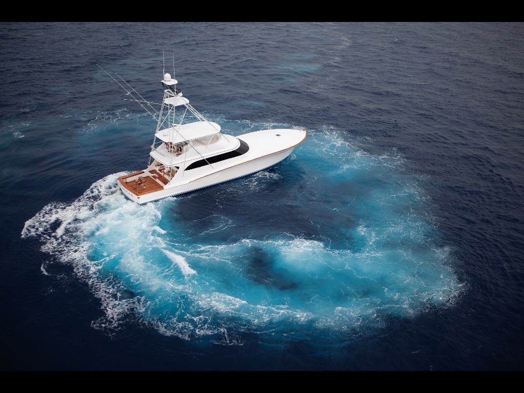 A motor yacht is seen cruising across deep blue waters, leaving a circular trail of foamy white wake. The sleek vessel's deck features wooden accents, and its white hull contrasts with the ocean's hues. The scene captures a tranquil yet dynamic maritime environment.