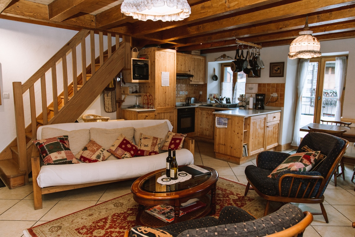 An open-plan living area features a comfortable sofa adorned with cushions, accompanied by two armchairs. A round coffee table sits at the center. Wooden cabinetry and appliances are clearly visible in the kitchen area, while a staircase leads to the upper level.