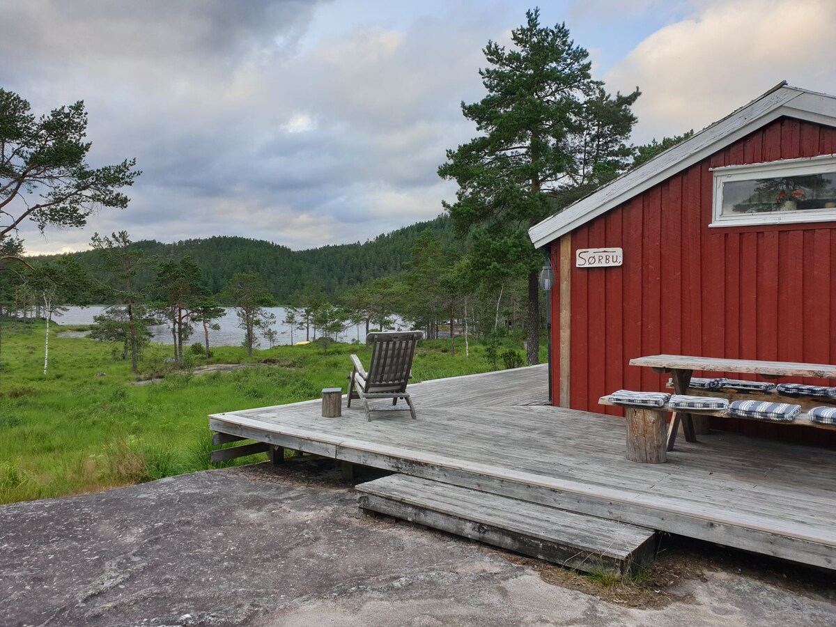 A rustic red cabin is positioned on a wooden deck, surrounded by an expansive grassy area and trees. A folding chair is placed on the deck, facing a serene body of water in the distance, with gentle hills beyond. Natural light enhances the tranquil scene.
