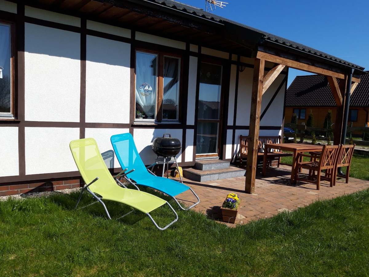 An outdoor area is visible featuring two reclining lounge chairs in bright green and blue. A wooden dining table with multiple chairs is positioned under a covered porch, complemented by a charcoal grill. Well-maintained grass surrounds the space, offering a relaxing environment.