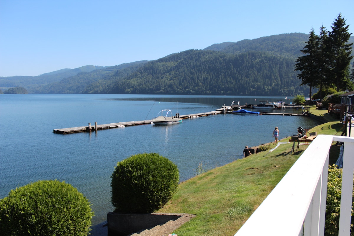 A serene lakeside view showcases a long dock extending into the calm waters of Lake Whatcom. Lush green hills frame the background, and a few small boats are visible along the shore. A well-maintained grassy area leads towards the water's edge.