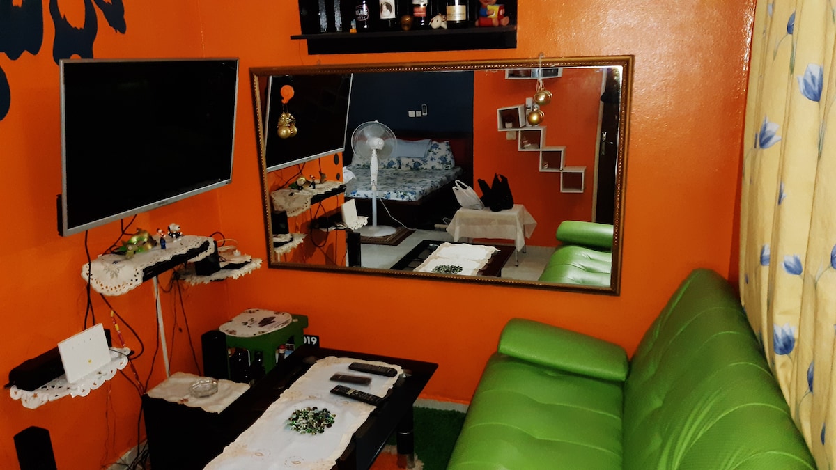 A vibrant living area features a green couch and a glass coffee table. A large mirror reflects the space, while a flat-screen TV is mounted on the wall. Soft lighting accentuates the orange walls, and a fan provides a cooling option. A glimpse of the bedroom area is visible in the background.