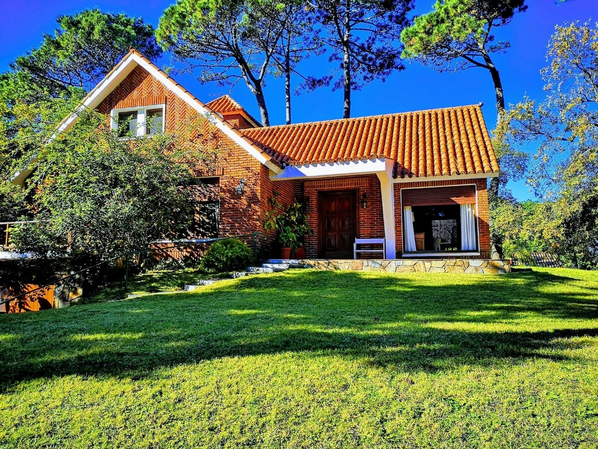 A charming chalet is surrounded by greenery, featuring a warm brick exterior and a traditional roof. A grassy lawn leads up to the entrance, where wooden doors invite exploration. Tall trees provide shade, enhancing the serene setting of this family-friendly retreat.