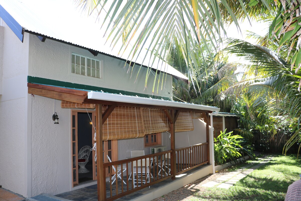 The exterior of the studio displays a wooden terrace with bamboo blinds that provide shade. Lush greenery surrounds the area, creating a serene environment. A small seating arrangement is visible on the terrace, inviting relaxation. Sunlight filters through palm leaves, enhancing the tranquil setting.