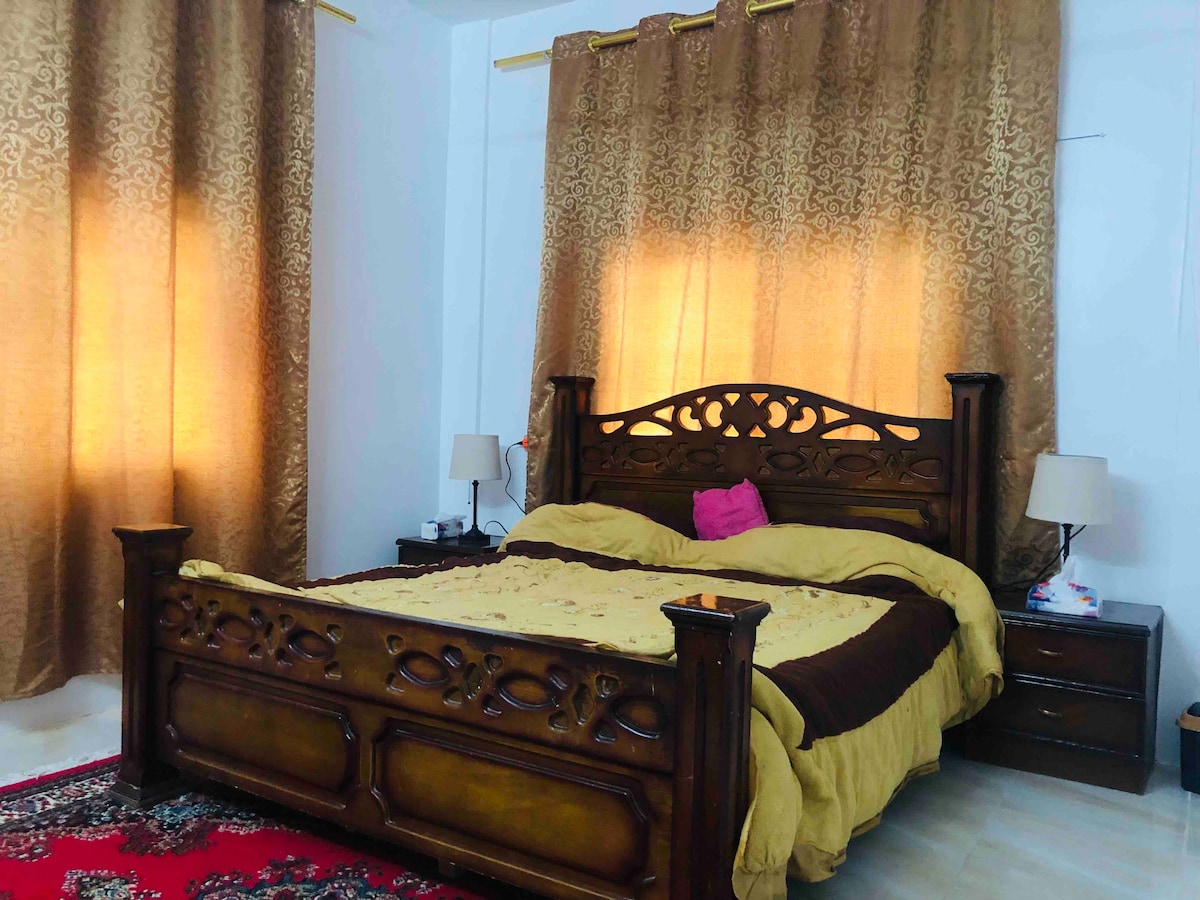 A spacious sleeping area features a wooden bed frame with an intricate design. Light brown curtains allow soft light to filter in, while two bedside tables hold lamps. A patterned rug adds a touch of warmth to the floor, complementing the overall decor.