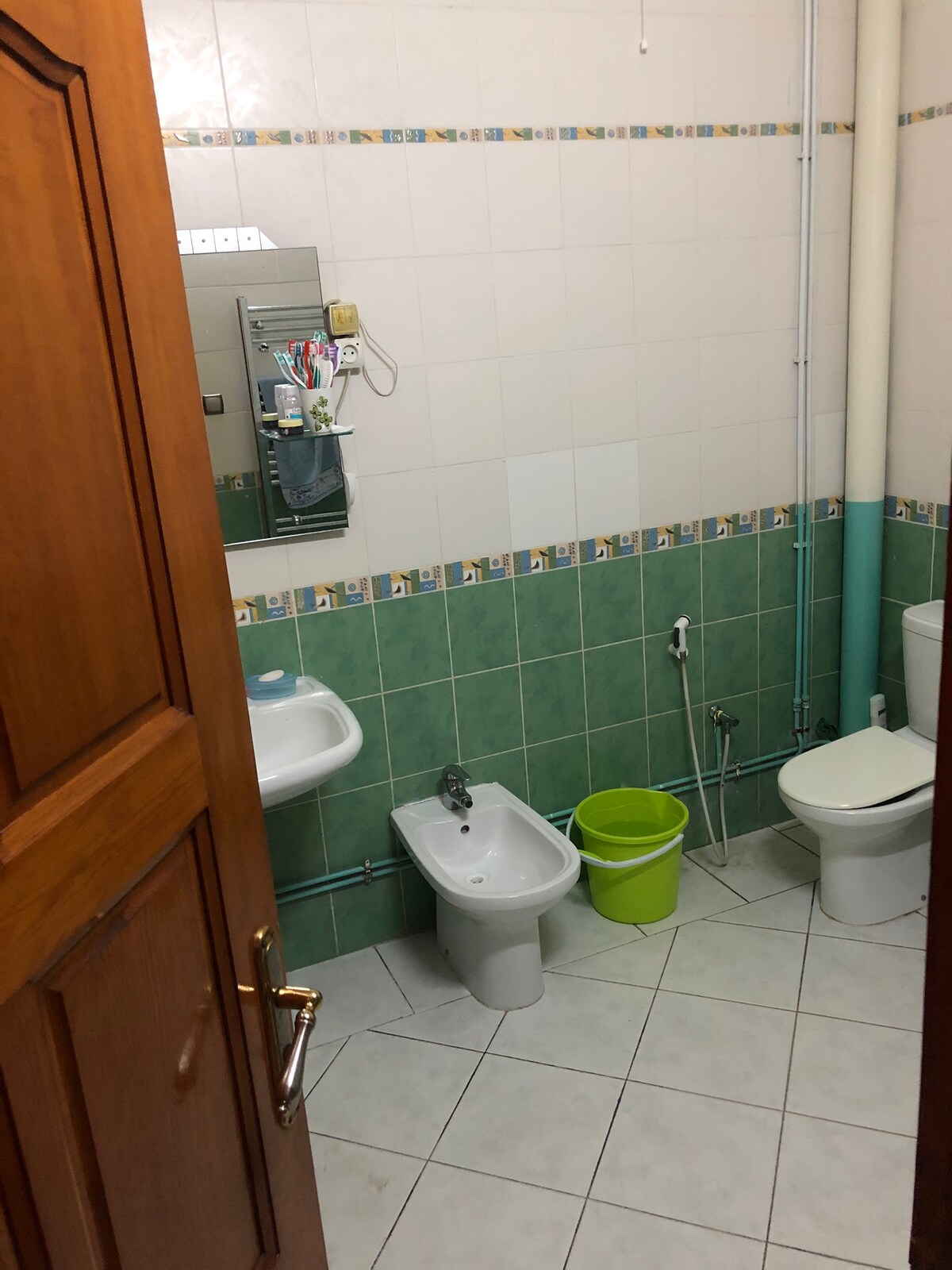 The bathroom is equipped with a toilet and a bidet, along with a small sink mounted on the wall. A mirror is positioned above the sink, and colorful storage items are visible nearby. The walls feature a green tiled design, and a yellow bucket is placed on the floor.