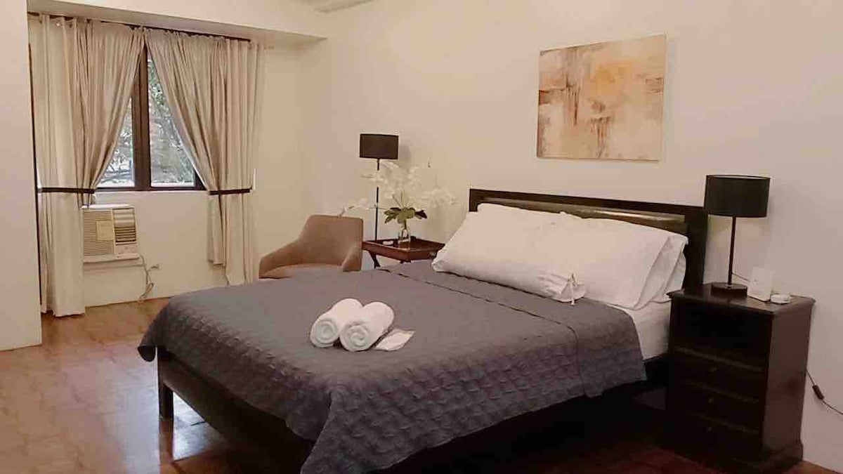 A well-lit bedroom features a spacious bed with neatly arranged white towels on top. A soft chair is positioned beside a nightstand, while a window with sheer curtains allows natural light to flow in. An air conditioning unit is visible, providing climate control.