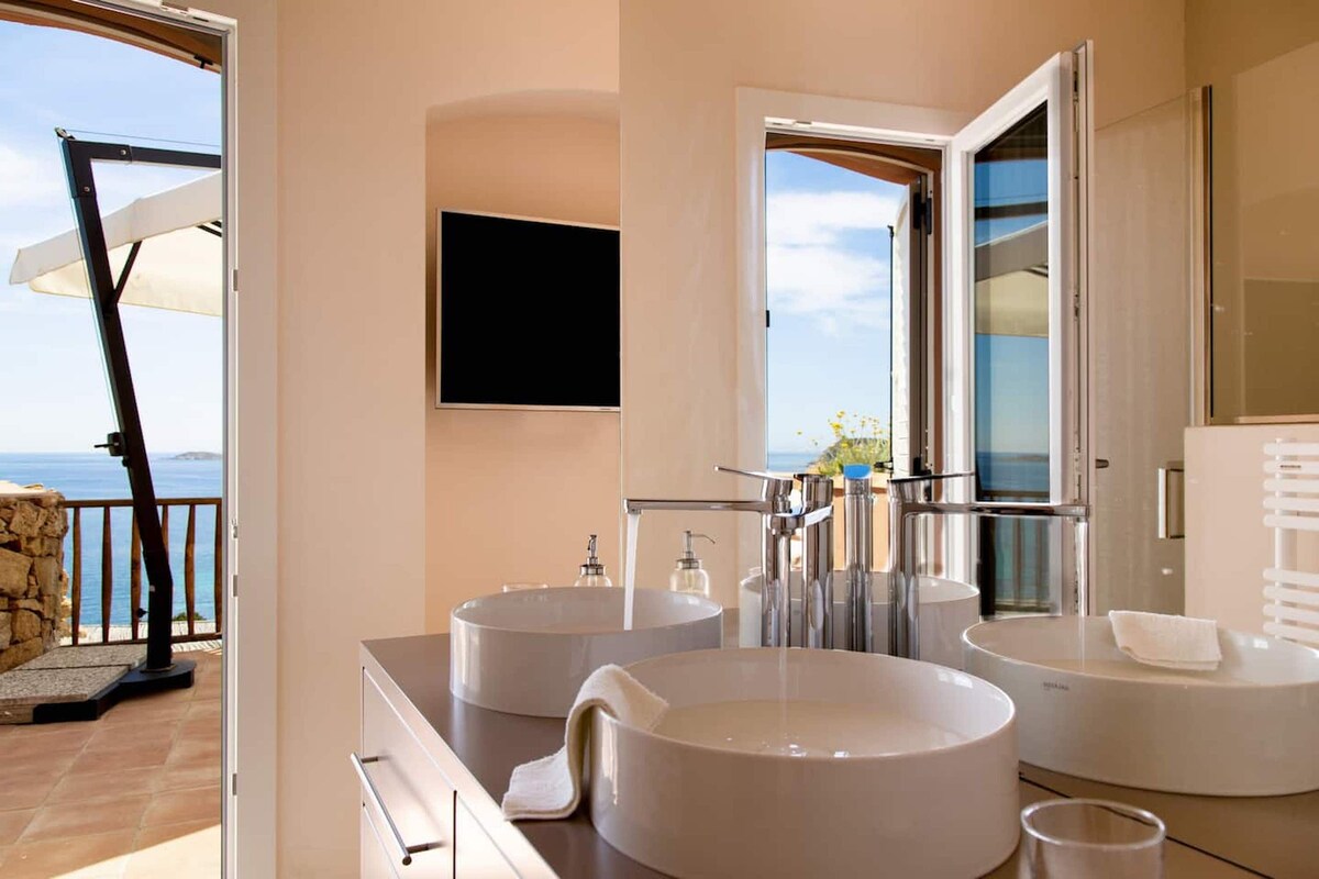 A well-appointed bathroom is presented, featuring dual round sinks on a spacious countertop. Large windows provide a view of the sea, while a wall-mounted television is visible. Natural light highlights the neutral tones of the room, enhancing the inviting atmosphere.