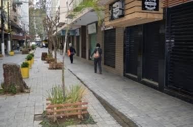 A tree-lined sidewalk is displayed with planters and paving stones. Storefronts are visible behind closed shutters. Two pedestrians walk down the pathway, accompanied by benches and greenery along the edges, contributing to a welcoming atmosphere in the urban setting.