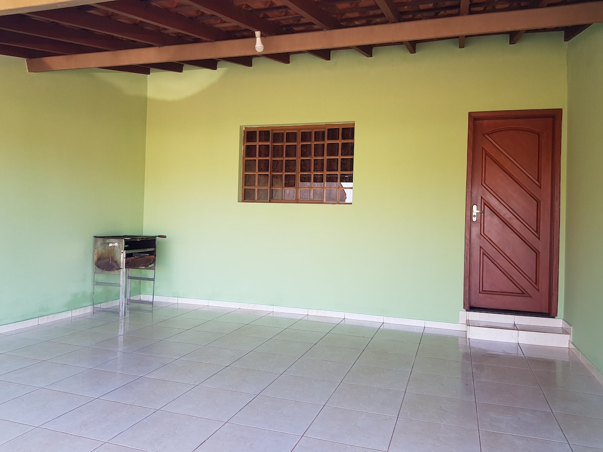 An open living area is presented with green walls and a tiled floor. A glass table is positioned against one wall, while a wooden door leads outside. Natural light enters through a grid-pane window, adding brightness to the spacious environment.