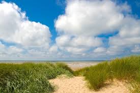 A sandy pathway is framed by lush green grass, leading toward the calm ocean. The blue sky is filled with fluffy white clouds, creating a serene coastal view that invites relaxation and exploration.