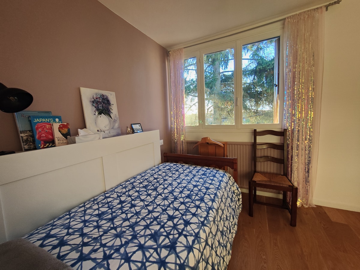 A private bedroom is presented with a bed featuring a patterned blue and white bedding set. Natural light enters through three windows, complemented by sheer curtains. A wooden chair and a small side table add functional elements, while art and books are arranged on a nearby surface.
