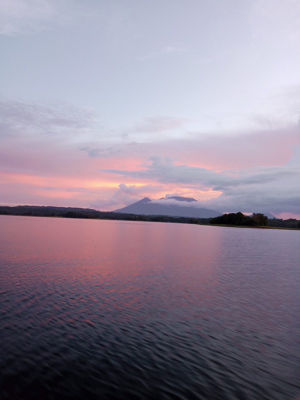 An expansive view of a serene lake captured at sunset, where soft pink and orange hues reflect on the water's surface. The backdrop features distant mountains under a partly cloudy sky, contributing to the tranquil atmosphere of the scene.