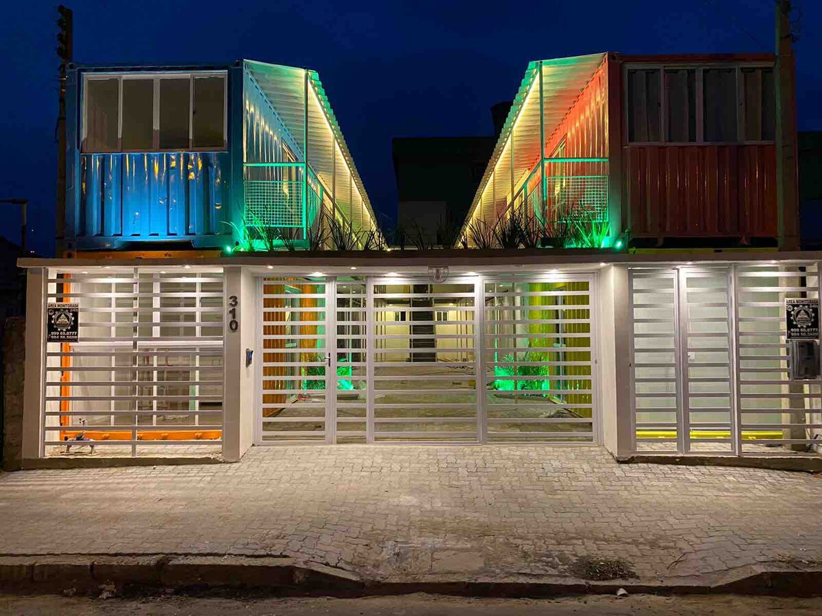 The exterior of the building showcases a modern architectural design featuring two container-style structures. Illuminated with colorful LED lights, the façade is framed by a grid of white fencing, providing a welcoming appearance in the evening setting.