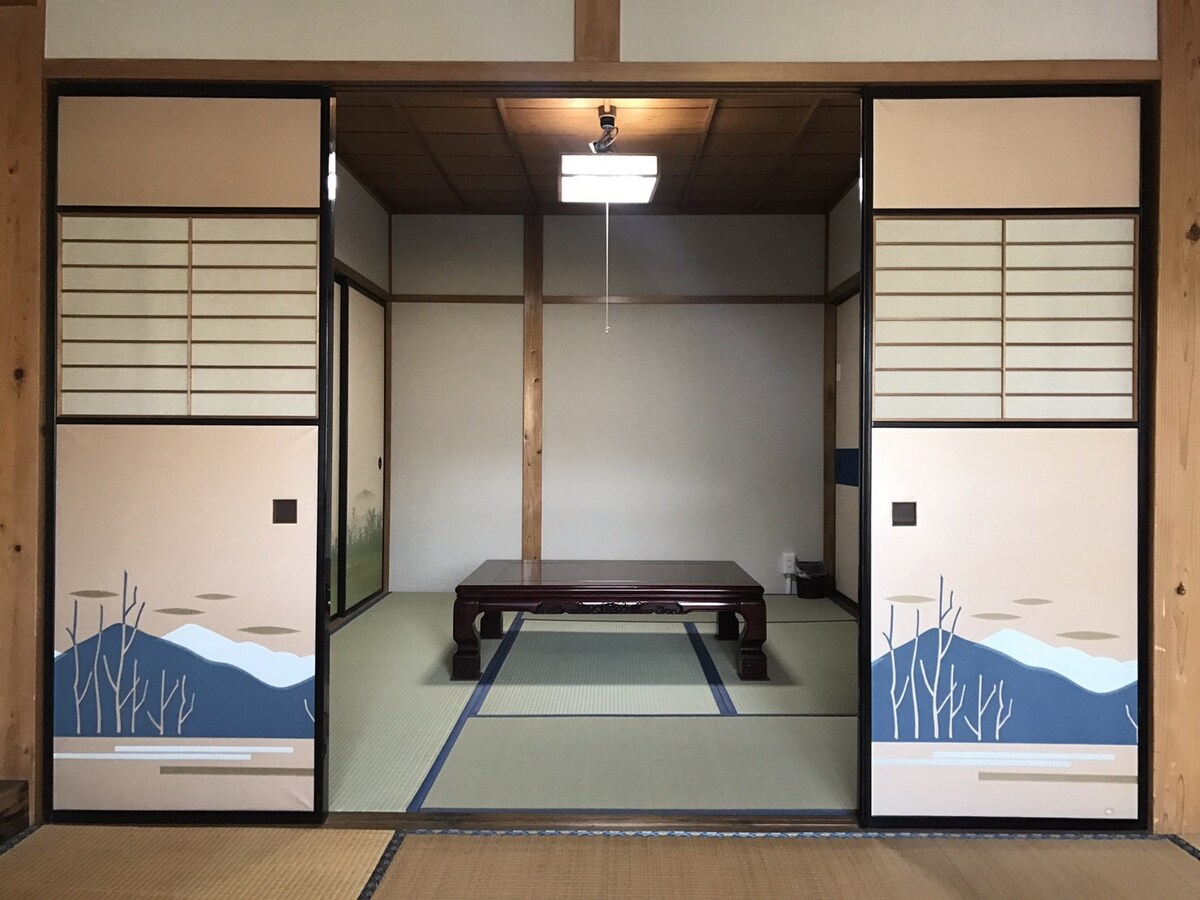 A traditional Japanese room is presented with sliding doors featuring subtle mountain designs. Tatami flooring is visible, complemented by a low wooden table at the center. Soft lighting enhances the serene atmosphere, while a glimpse of a natural scene is observed through the doors.