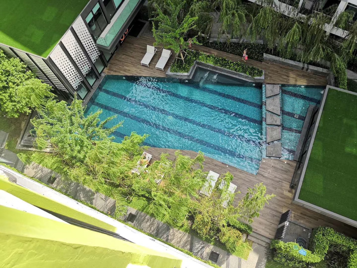 An aerial view of the community pool is presented, surrounded by well-maintained greenery and wooden decking. The pool features a clear blue water surface, with several lounge chairs positioned nearby for relaxation.