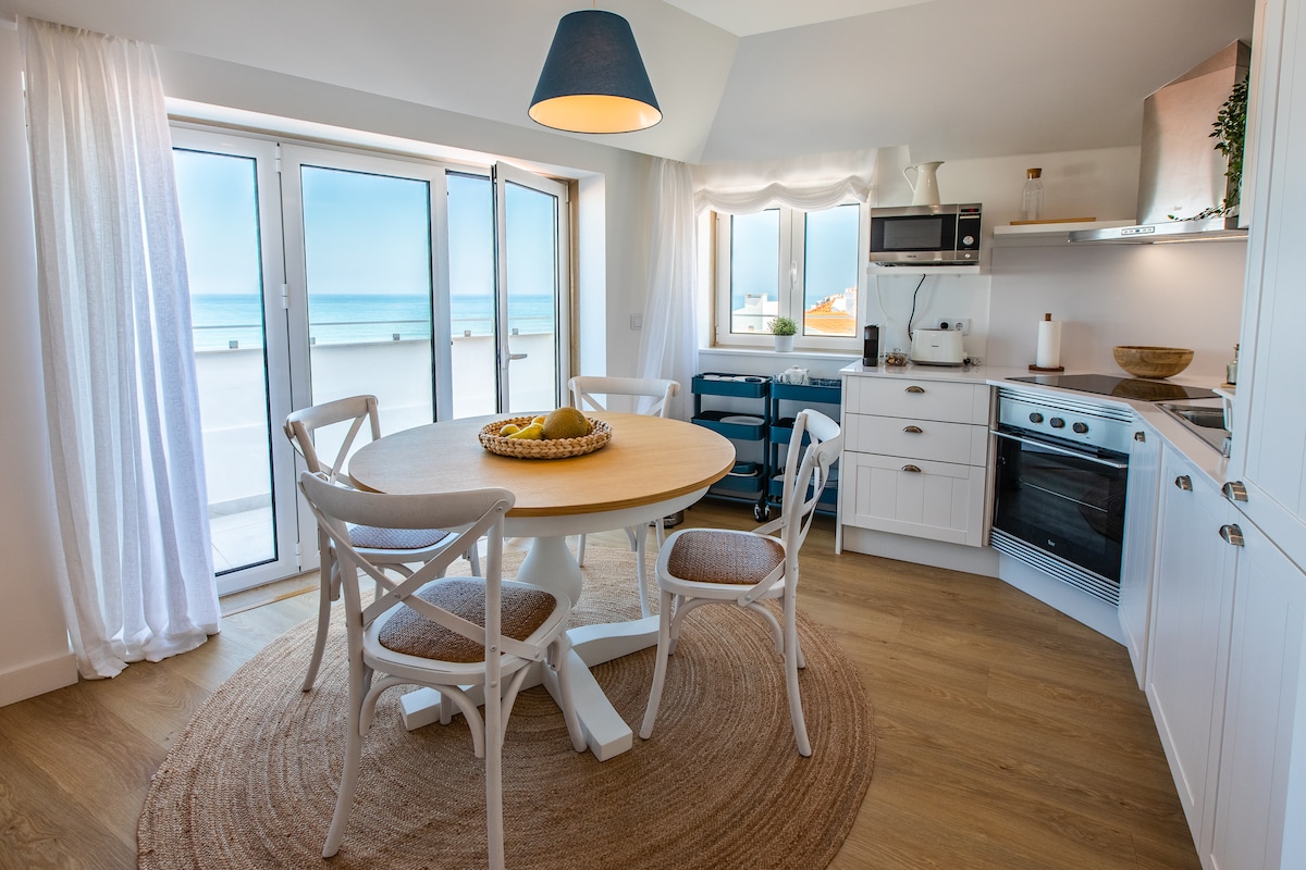 A bright kitchen space features a round dining table surrounded by four chairs. The kitchen is equipped with modern appliances and cabinetry, with natural light streaming in through large sliding glass doors that lead to a terrace with views of the ocean.