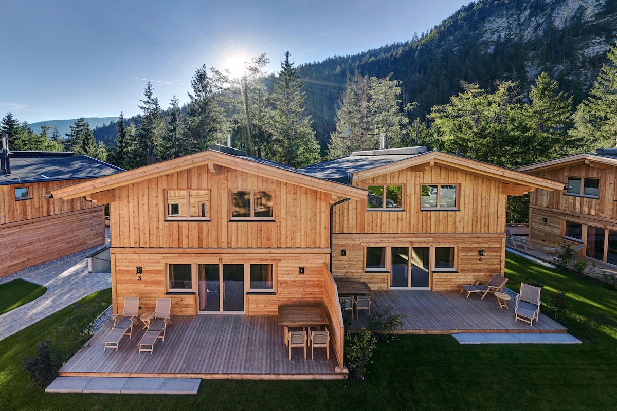 A wooden lodge is set among trees and mountains, featuring large windows that allow natural light to fill the interior. Outdoor seating is arranged on a wooden deck, providing space for relaxation and enjoyment of the surrounding nature.