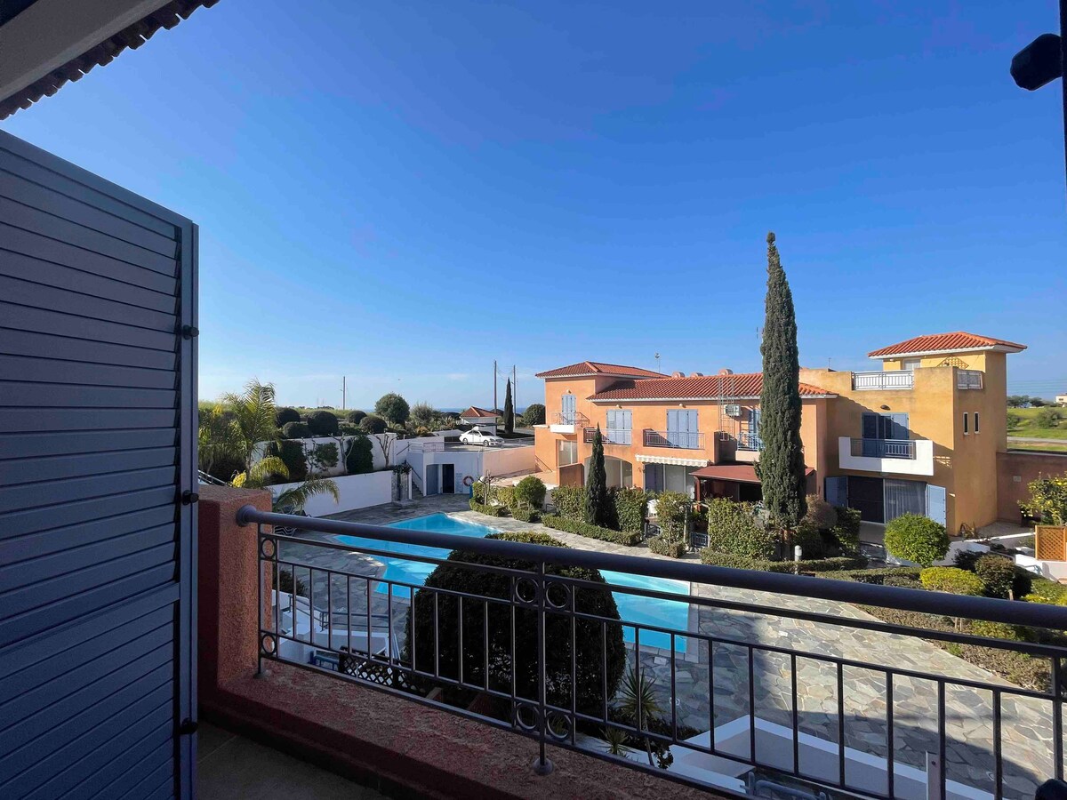 A balcony view captures the serene landscape, featuring a communal pool surrounded by well-maintained gardens. The image showcases a peaceful residential complex with clear skies and distant greenery, creating a tranquil backdrop for relaxation.
