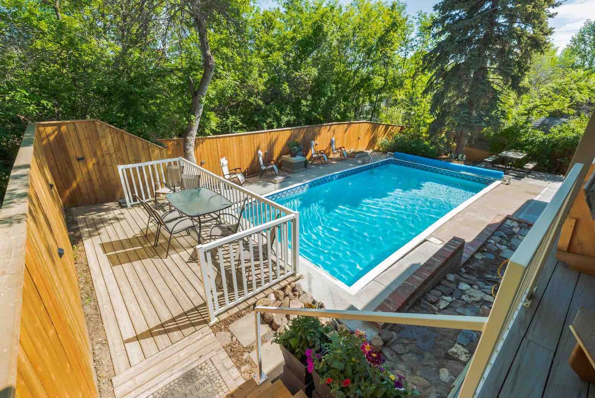 An aerial view of a spacious outdoor area features a rectangular in-ground pool surrounded by a wooden deck. Several lounge chairs are positioned along the poolside, and a table set with chairs is visible nearby. Lush greenery provides a natural border around the space.