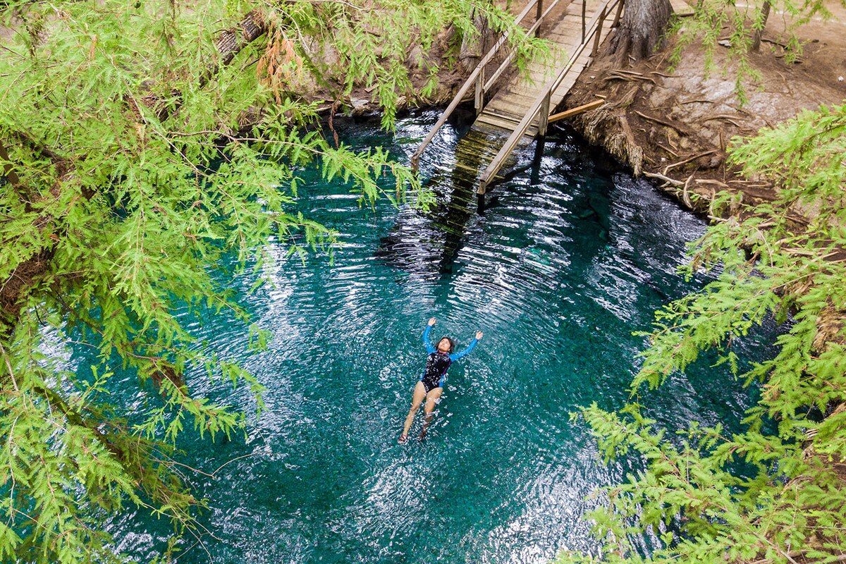 A tranquil swimming area is surrounded by lush greenery, showcasing clear blue waters. A wooden bridge spans across the water, connecting both sides of the natural setting. A person is seen floating peacefully, enjoying the serene environment.