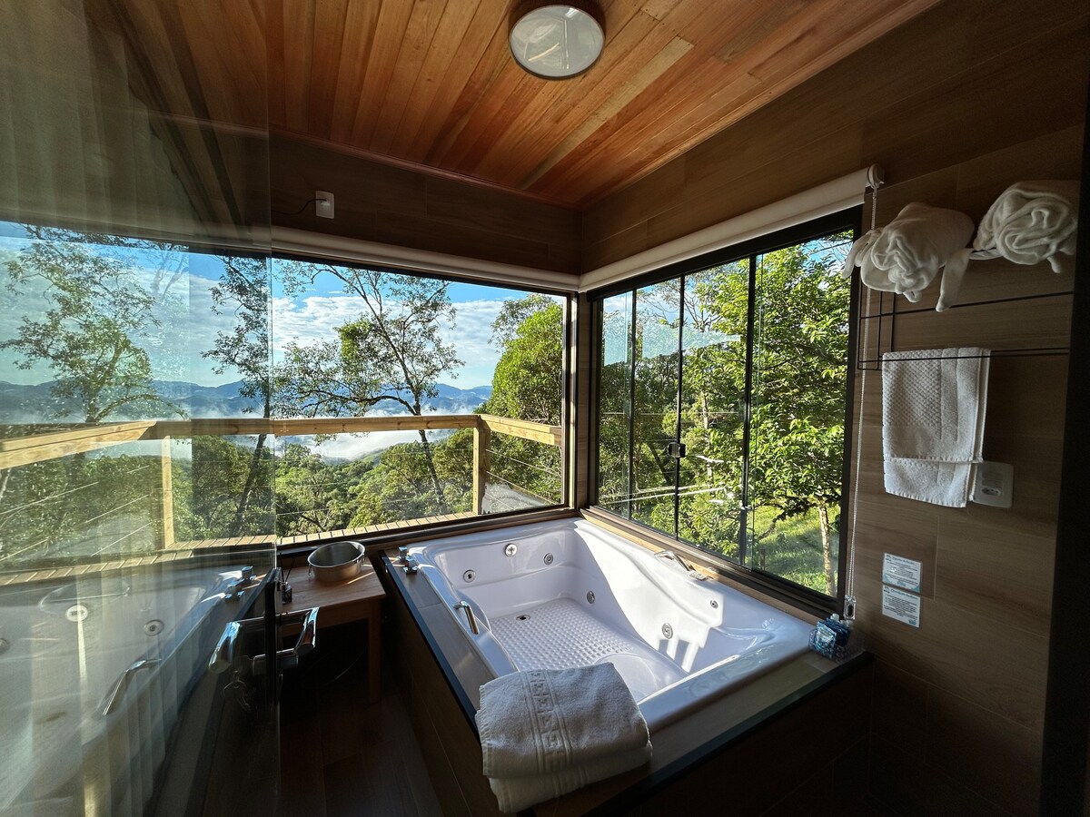 A modern bathroom features a spacious hot tub positioned against a large window, offering views of surrounding greenery and mountains. The wooden ceiling adds warmth, and soft towels are neatly arranged nearby. Natural light floods the space, enhancing the serene atmosphere.