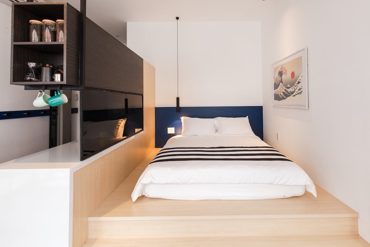 A minimalist studio layout features a platform bed with a striped duvet, surrounded by sleek wooden surfaces. A kitchenette is visible, equipped with glass jars and coffee-making tools. The wall is adorned with a serene artwork depicting a sunrise over a wave.