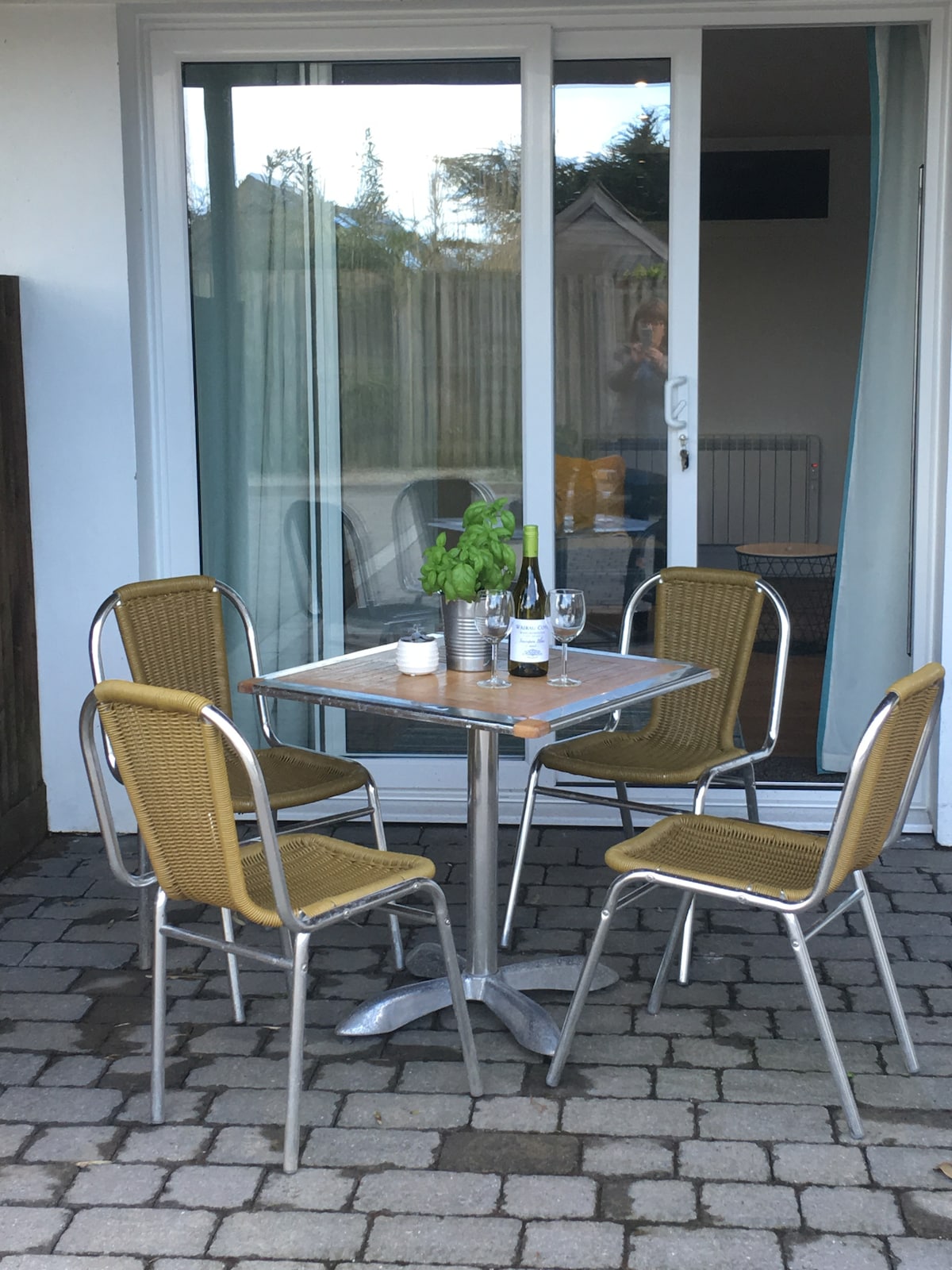 An outdoor patio area features a small table surrounded by four metal-framed chairs with woven seats. The table holds two wine glasses and a bottle of wine, complemented by a potted plant. Large glass doors provide access to the interior.