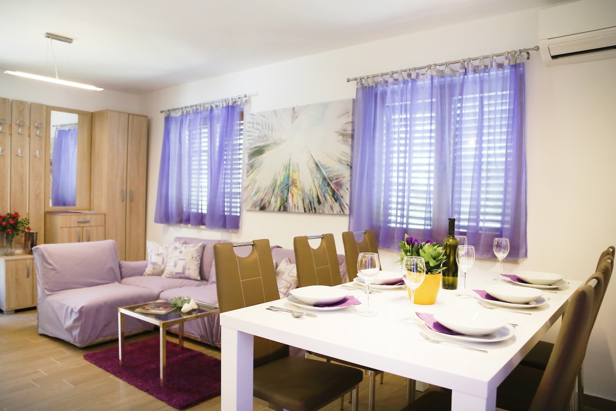 A dining area features a white table set for six, with plates and glasses arranged for a meal. A cozy couch in soft tones provides additional seating. Large windows with sheer purple curtains allow natural light to fill the space, complemented by modern artwork on the wall.