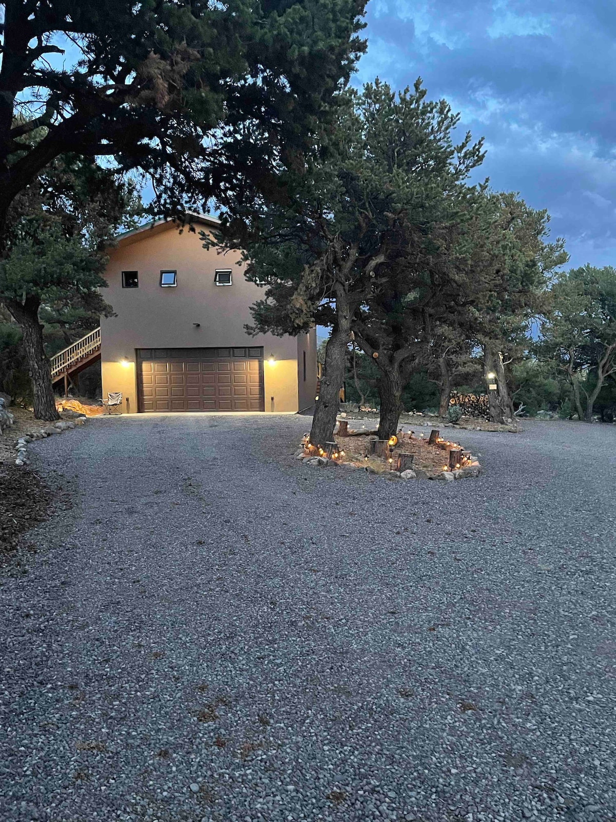 A newly constructed carriage house is set amid trees, featuring a modern facade with subtle lighting. A gravel driveway leads to the garage, while a gathering space surrounded by stones and lights is seen in the foreground, inviting relaxation in a serene environment.