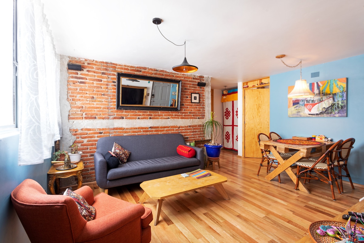 A vibrant living area showcases exposed brick walls paired with light blue accents. A comfortable gray sofa is positioned next to a wooden coffee table. A cozy red chair complements the space, and a dining area is visible with a wooden table surrounded by elegant chairs.