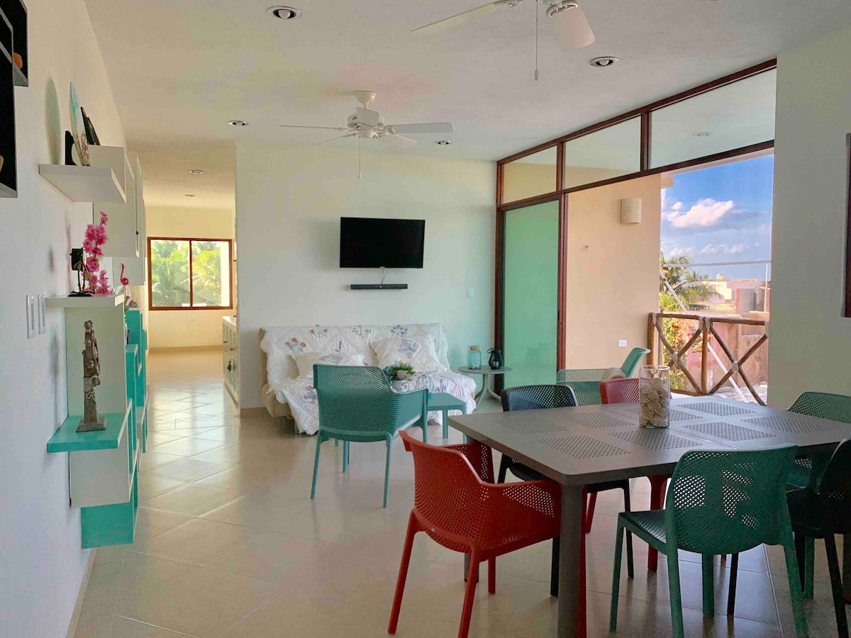 An open living and dining area features a comfortable sofa and a dining table surrounded by colorful chairs. A large window offers natural light and views of the surroundings. A wall-mounted television is positioned for easy viewing, and light tiles accentuate the space.