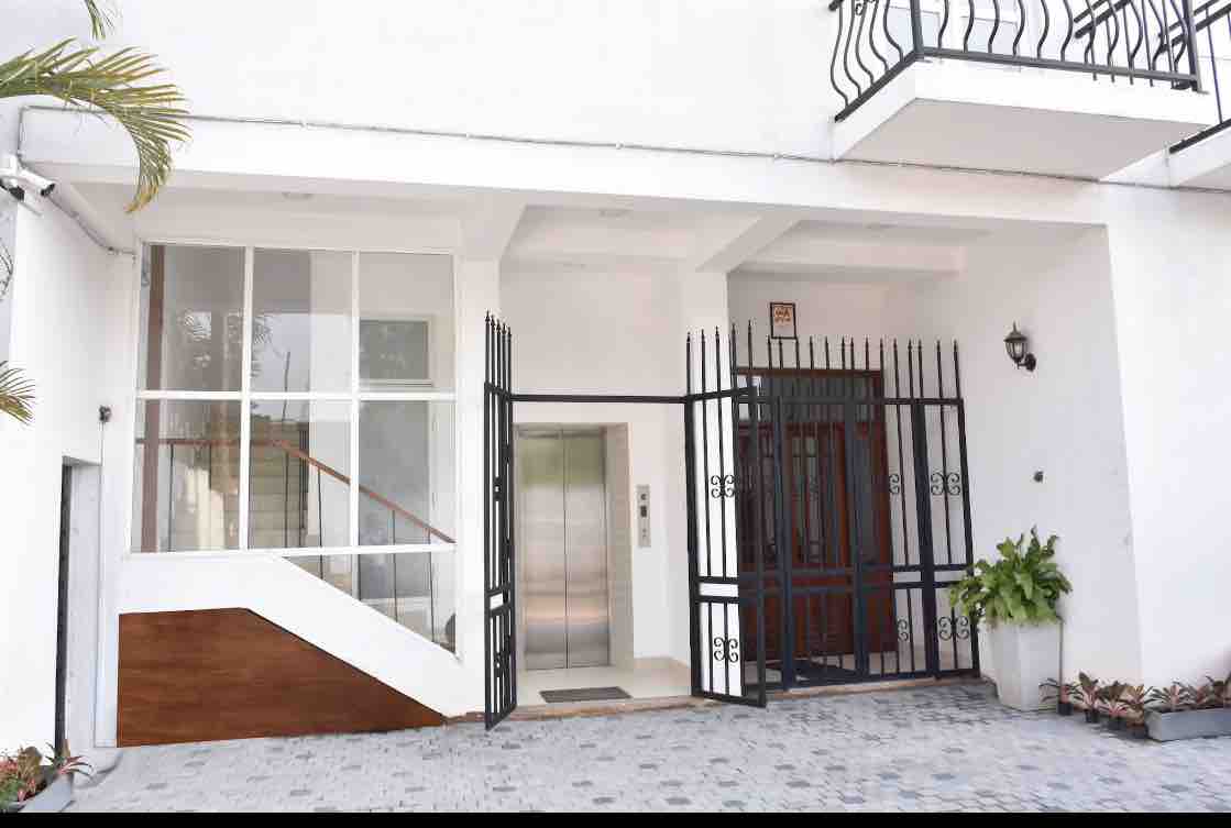 The entrance to the apartment building features a secure gate and an elevator, providing convenient access. Large windows allow natural light to illuminate the space. Potted plants are arranged near the entrance, enhancing the welcoming look of the exterior.