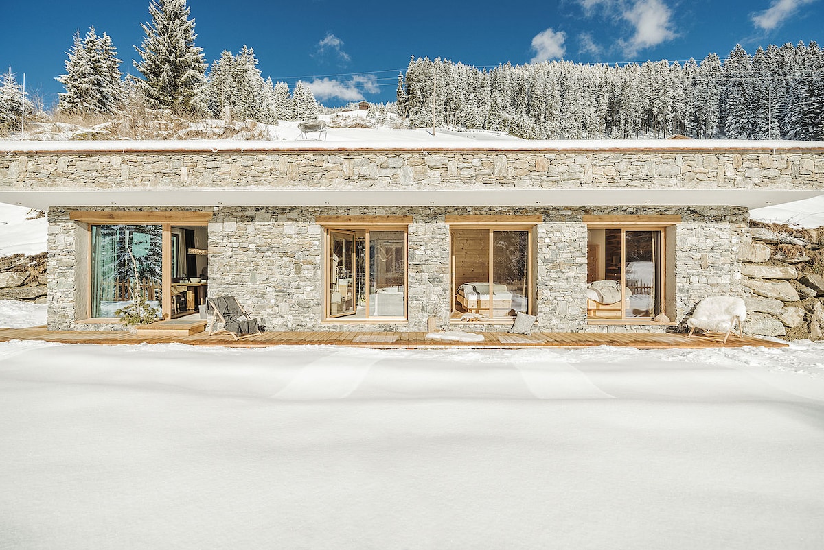 A modern stone structure is nestled in a snowy landscape. Large glass windows offer views of the serene surroundings, including evergreen trees. The entrance features a covered patio area, contrasting the white snow with natural stone textures.