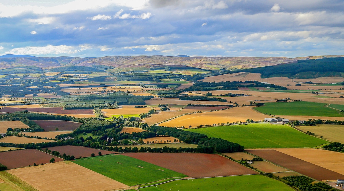 An expansive view of the Scottish countryside illustrates rolling hills and fields in various shades of green and brown. The landscape is dotted with patches of trees, and distant hills create a gentle horizon under a partly cloudy sky.