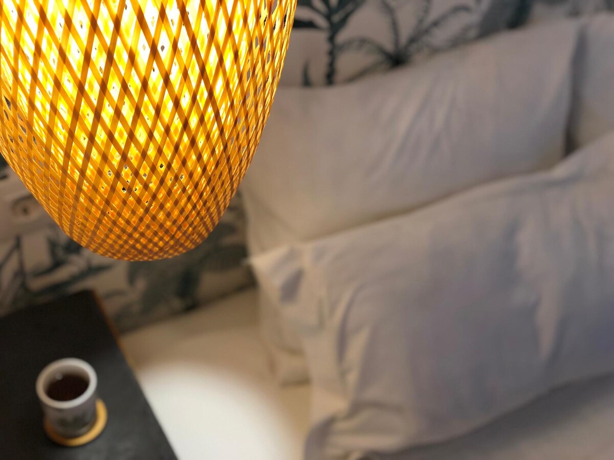 A cozy sleeping area is illuminated by a warm pendant light overhead. The spacious bed is adorned with fluffy white pillows, complemented by a bedside table featuring a small decorative item. The backdrop reveals a patterned wall, enhancing the room's tranquil ambiance.