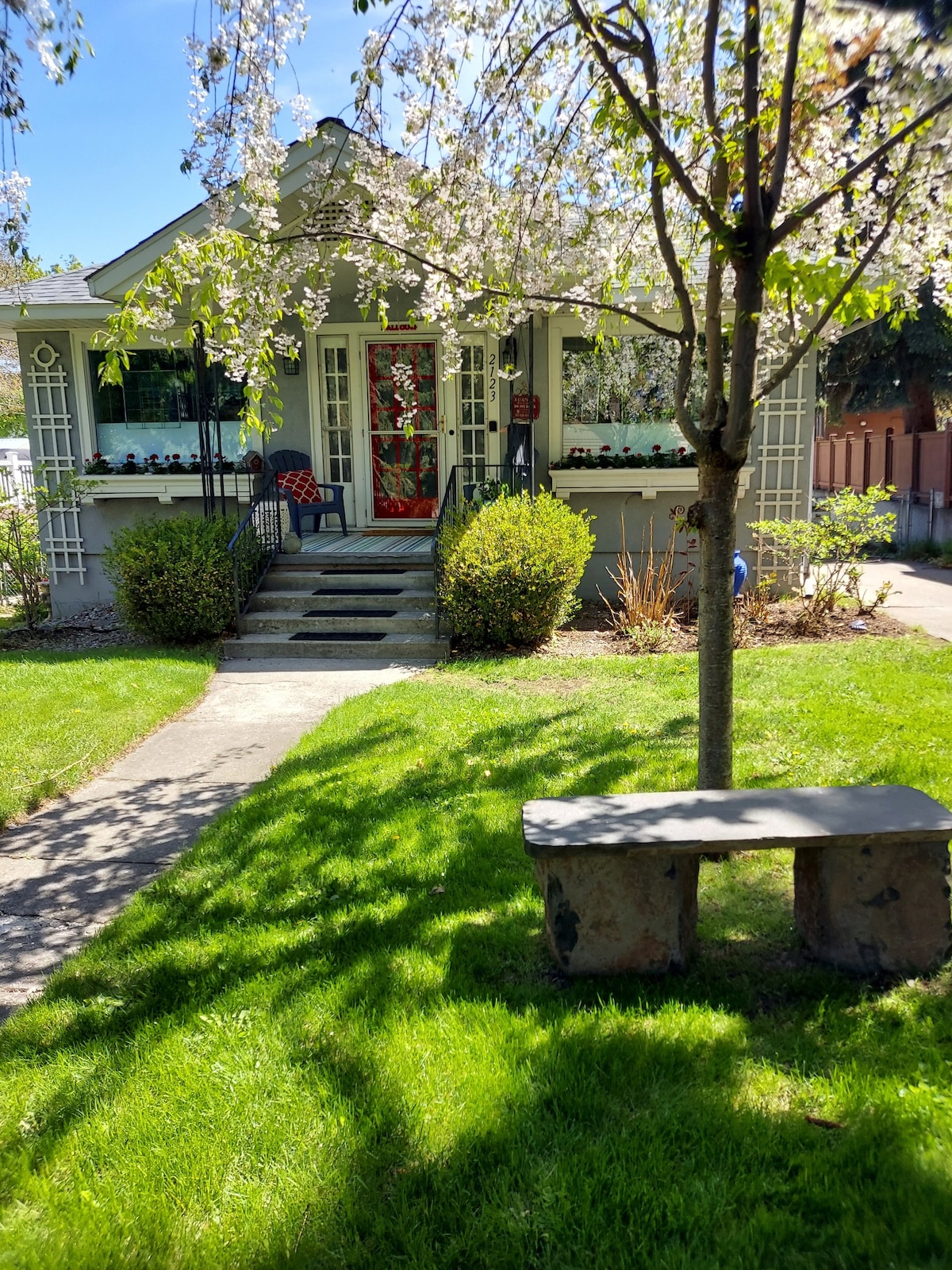The exterior of a charming house is framed by blooming trees, with a well-maintained lawn and pathway leading to a red door. Flower pots adorn the porch, and a rustic stone bench sits under the shade of a nearby tree.