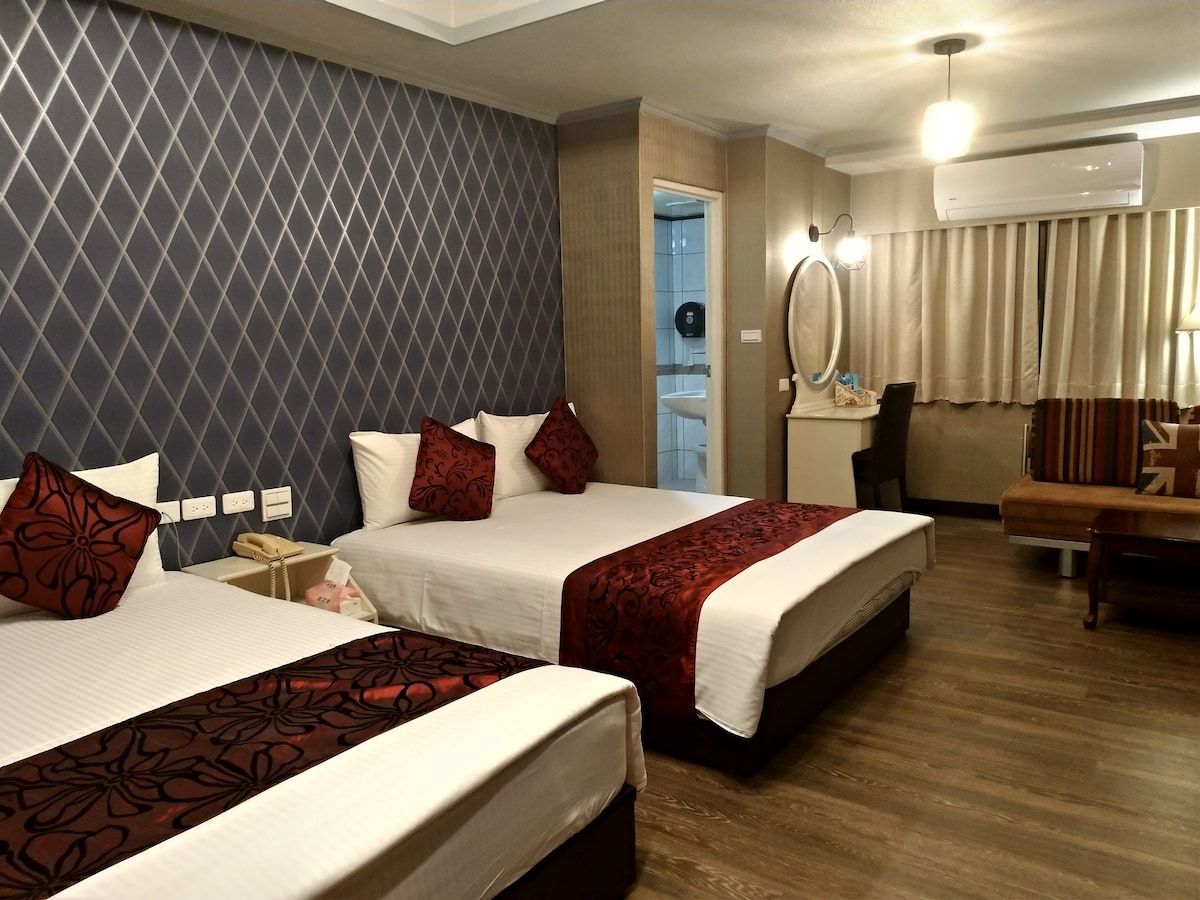 The room features two spacious beds adorned with decorative red and dark patterned throws. A seating area with a brown sofa is visible, along with a large mirror and a desk. A private bathroom entrance can be seen in the background.