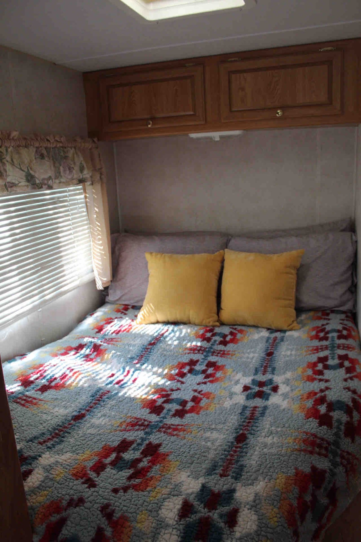 A cozy sleeping area is featured with a double bed covered by a colorful blanket in red, blue, and yellow hues. Two yellow pillows rest against the headboard, and natural light enters through a window adorned with light fabric blinds.