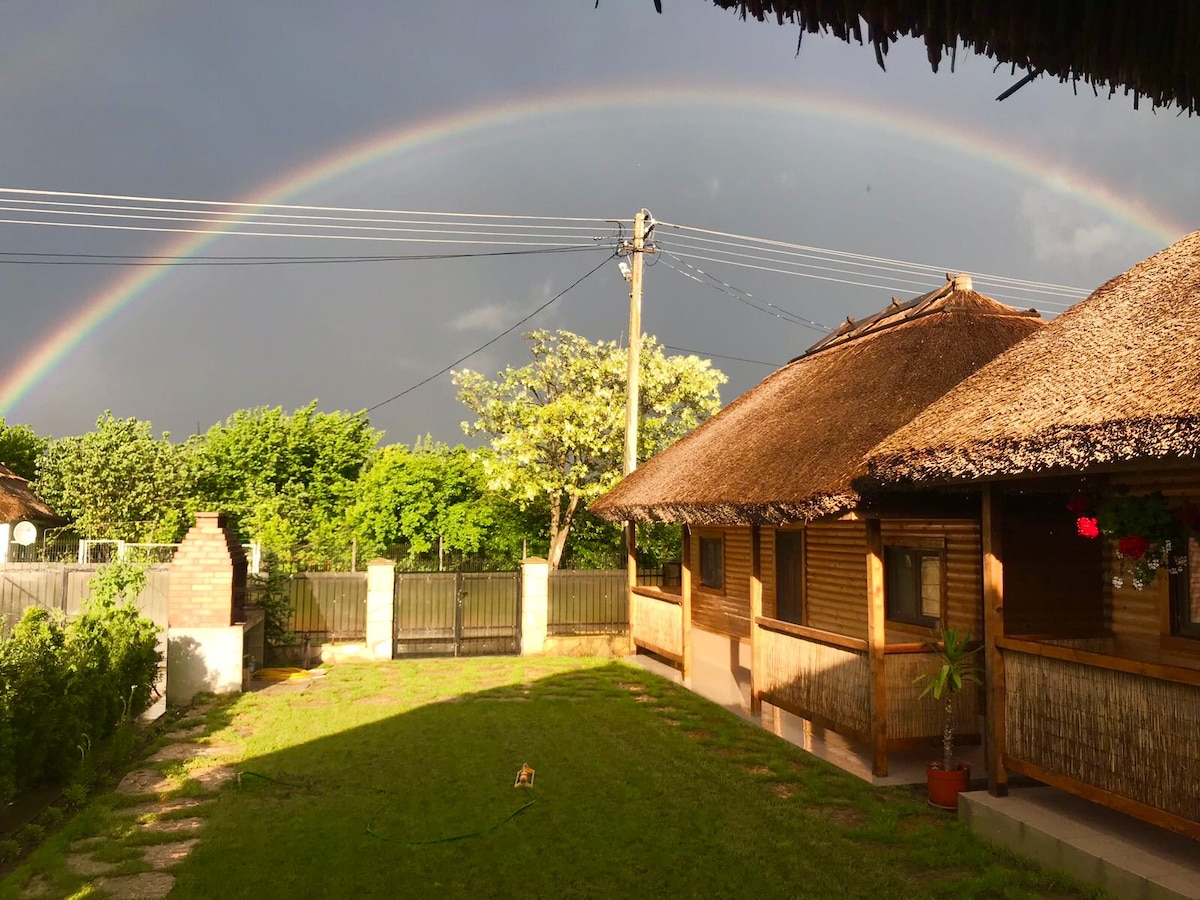 A vibrant rainbow arches over the property, framed by lush greenery. Traditional wooden houses with reed roofs are visible on either side, situated in a spacious courtyard with a well-maintained lawn. Nearby, a stone pathway leads towards a gated entrance.