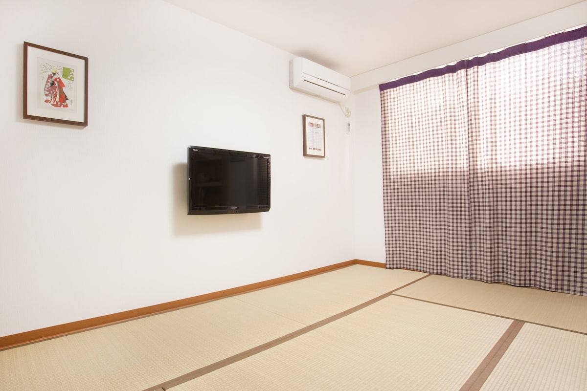 A bright room features tatami flooring and is complemented by a flat-screen TV mounted on the wall. Natural light enters through a window adorned with checkered curtains, with air conditioning visible on the wall. Simple wall art adds a touch of character.