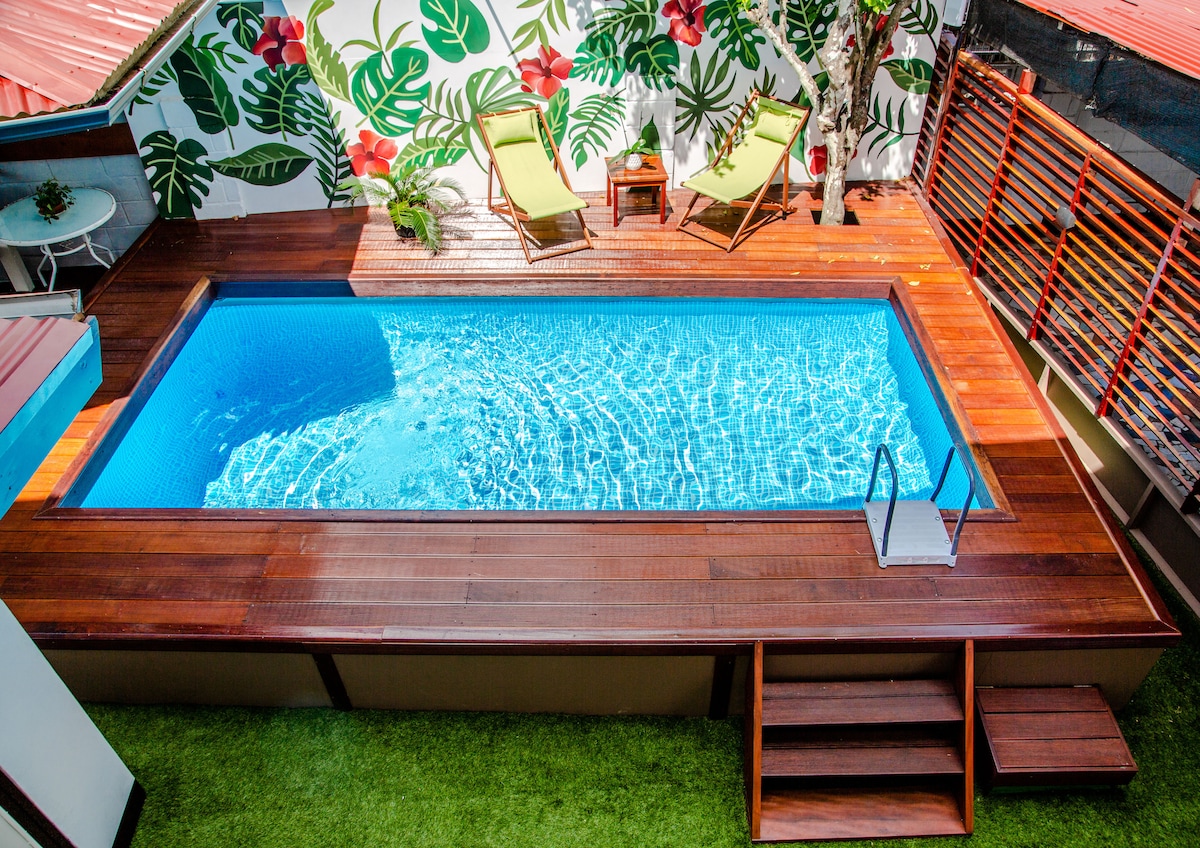 A rectangular pool is surrounded by a wooden deck, showcasing sparkling blue water. Tropical-themed murals with large green leaves and red flowers provide a vibrant backdrop. Two lounge chairs are positioned beside the pool, offering a relaxing space. Steps lead into the pool area from the deck.