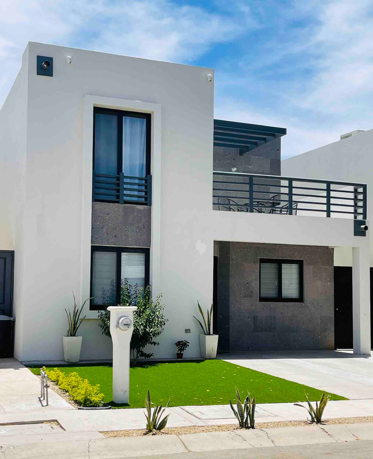 The exterior of the modern residence is characterized by a sleek façade and large windows. A small patch of green grass and decorative plants adorn the front yard, adding a touch of nature. A balcony extends from the upper level, providing outdoor space.