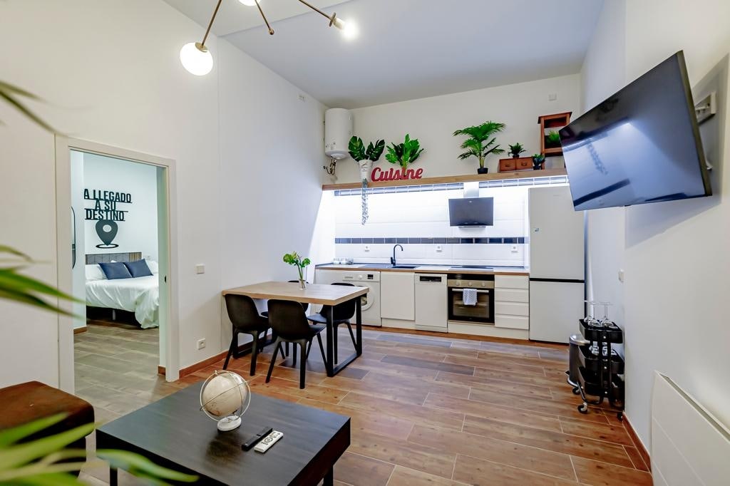 An open-concept living area features a kitchen equipped with modern appliances and a dining table for four. A comfortable sofa and a wall-mounted TV enhance the space. Plants add a touch of greenery, while a glimpse of a separate bedroom is visible through the doorway.