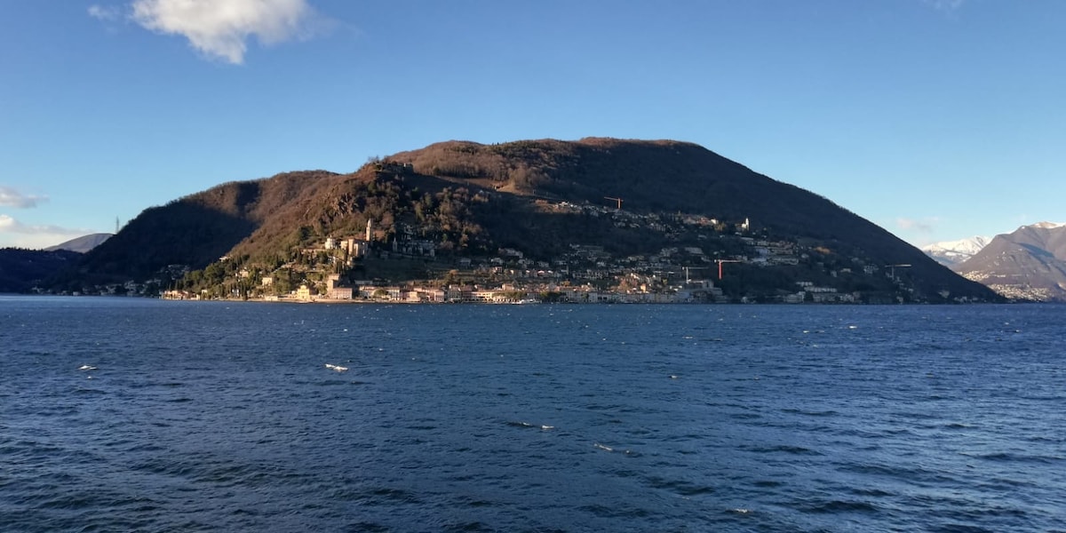 The image captures a prominent view of a hillside village along the edge of a lake, surrounded by mountains. The landscape features a mix of greenery and structures, with a clear blue sky above and gentle ripples on the water's surface.