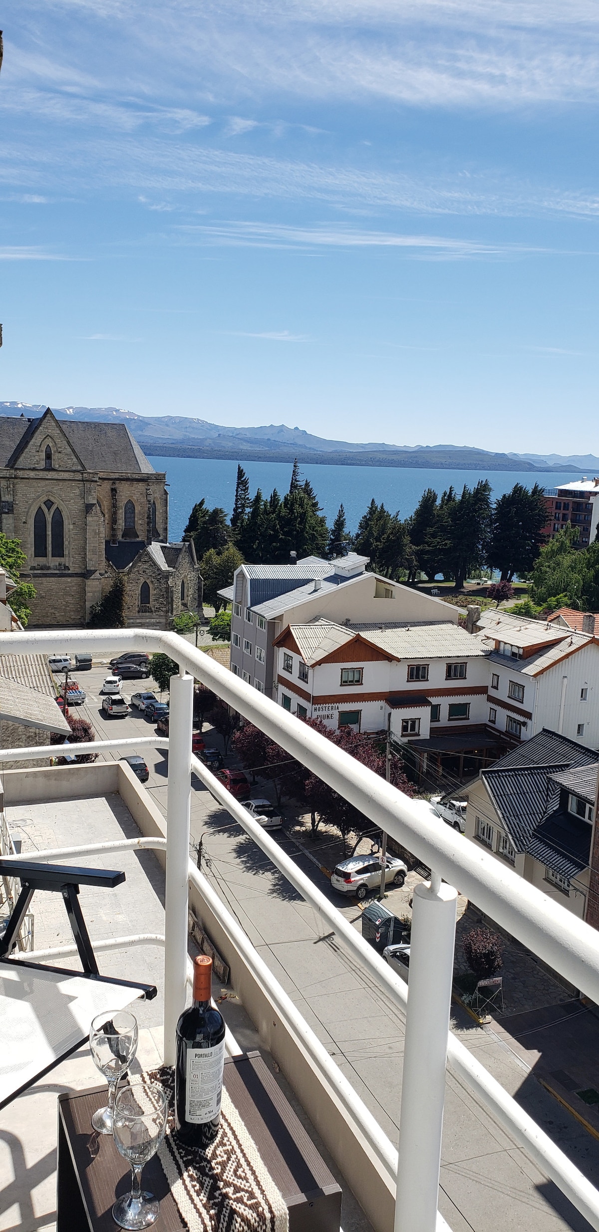 A balcony showcases a scenic view of the lake and surrounding buildings, along with a glimpse of the nearby church. A small table is set with two glasses and a bottle of wine, inviting guests to enjoy the outdoor space.