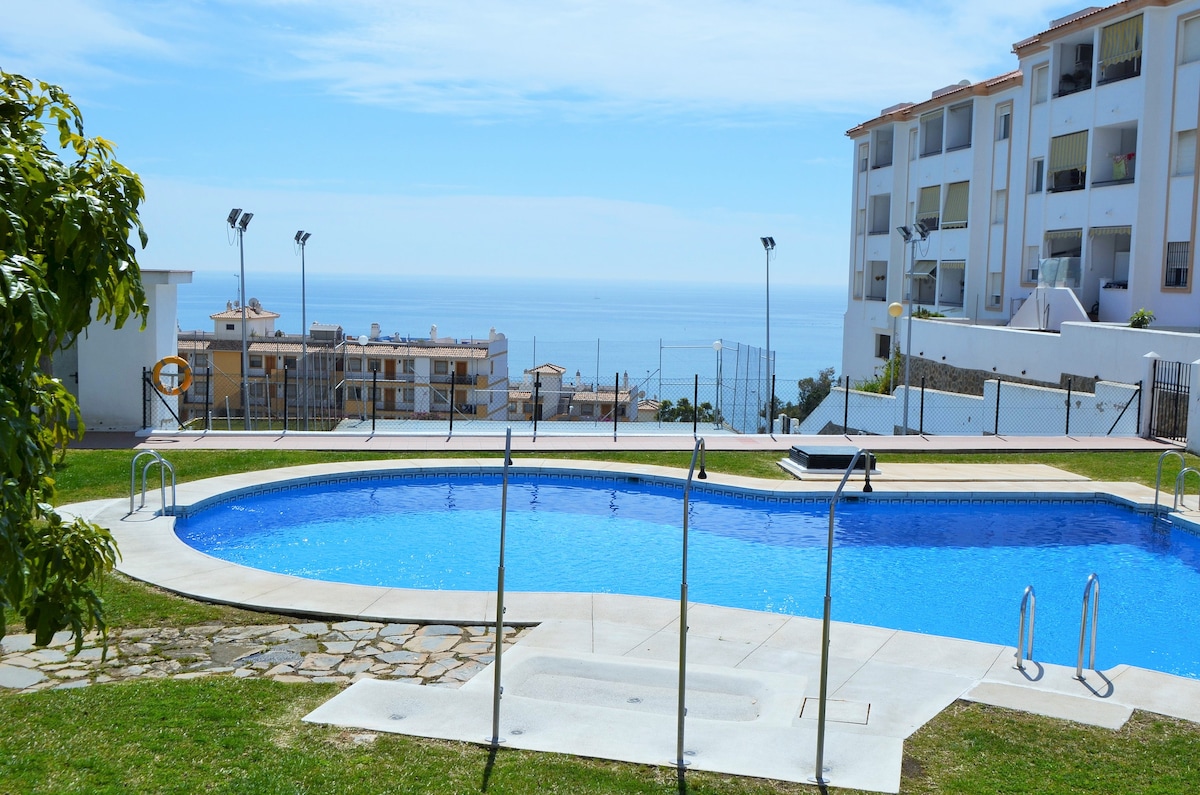 A clear blue swimming pool is surrounded by green grass and framed by a modern residential building. The ocean is visible in the background, providing a serene backdrop. The area includes poolside loungers and a spacious deck, enhancing the outdoor leisure space.