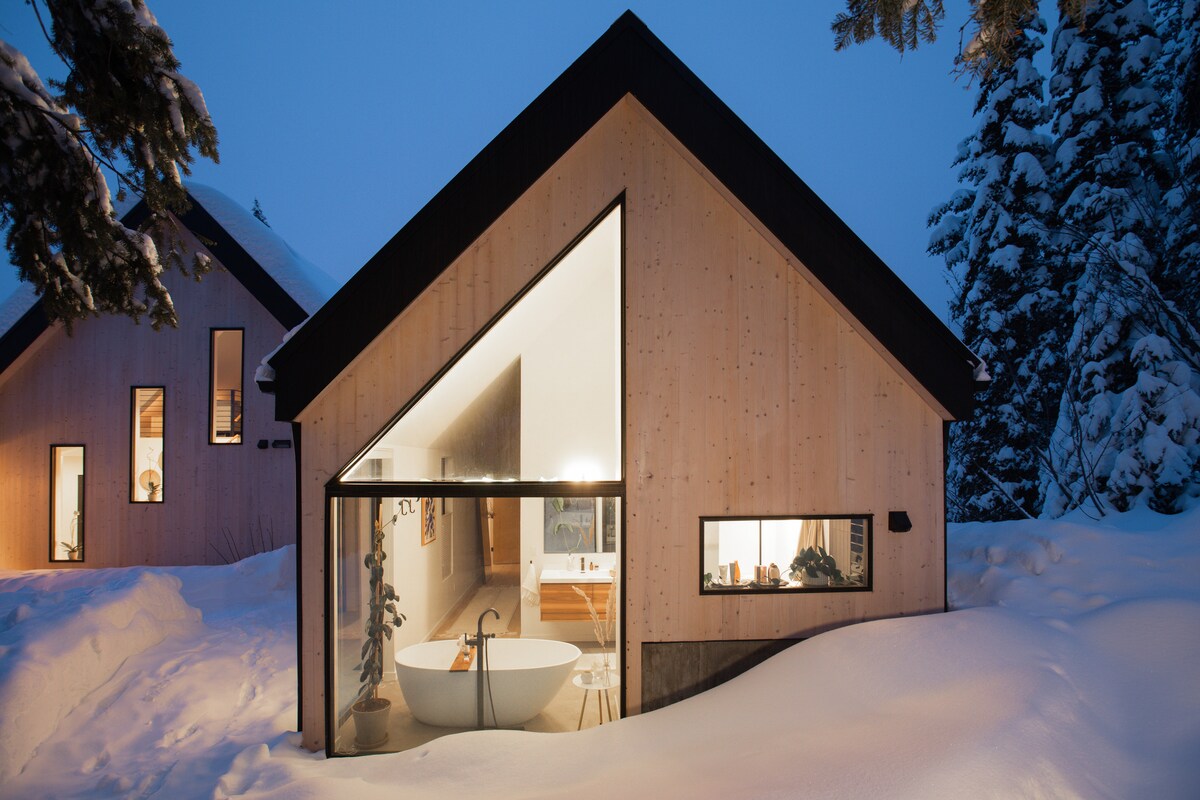 The modern exterior of the retreat is showcased under a winter sky, featuring a unique triangular design. A large glass wall reveals a vibrant bathroom space with a standalone tub, surrounded by snow-covered landscape, emphasizing the harmony between indoor and outdoor elements.