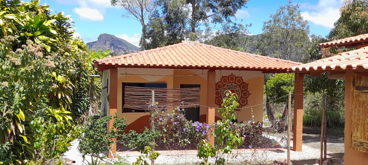 A charming single-story chalet is surrounded by lush greenery, featuring a terracotta roof and colorful mural on one side. The inviting outdoor space includes a variety of plants, while the background showcases rolling hills under a bright blue sky.