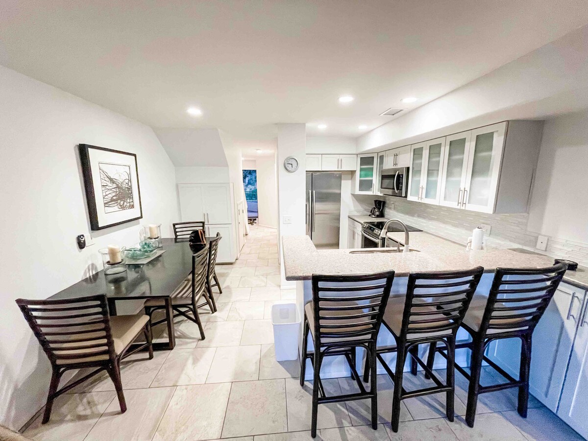 A modern kitchen area is displayed with sleek cabinetry and a spacious countertop. Barstools line the island, providing seating for four. A dining table is positioned nearby, equipped with chairs and tasteful decor. Large appliances including a refrigerator and coffee maker are visible.